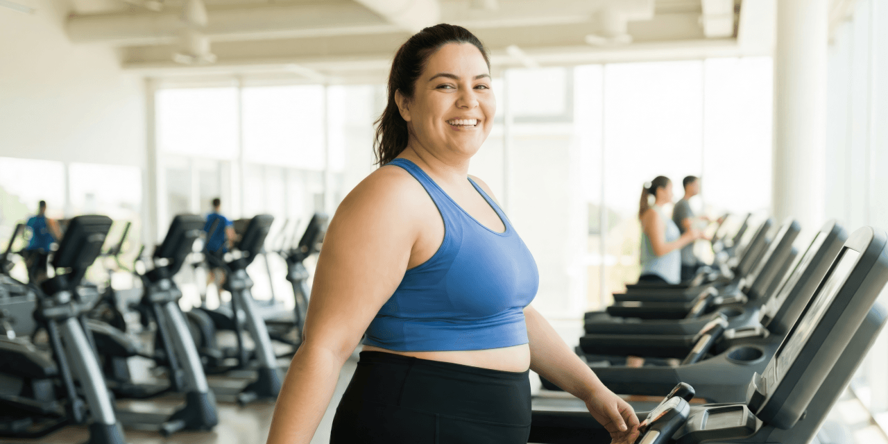 Young woman smiling while walking on treadmill in a modern gym. She's wearing a deep blue sports bra and black leggings.