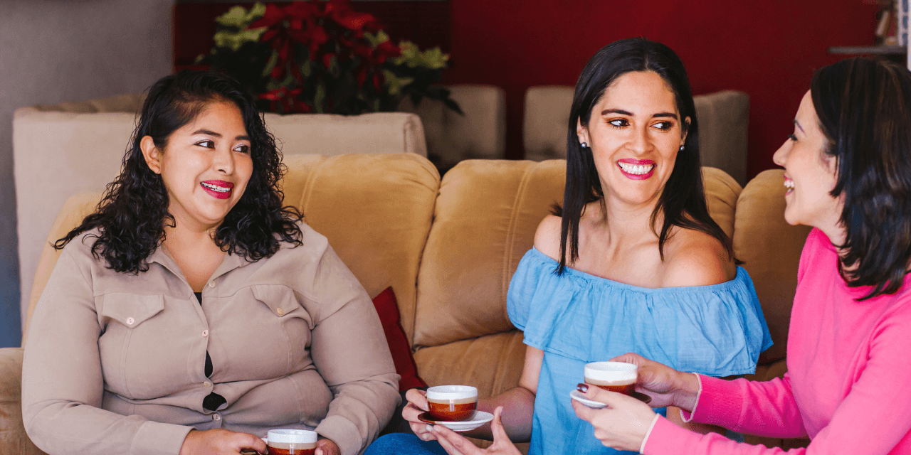 Three women chatting, offering support to each other. They're sitting on a sofa in a cozy cafe.