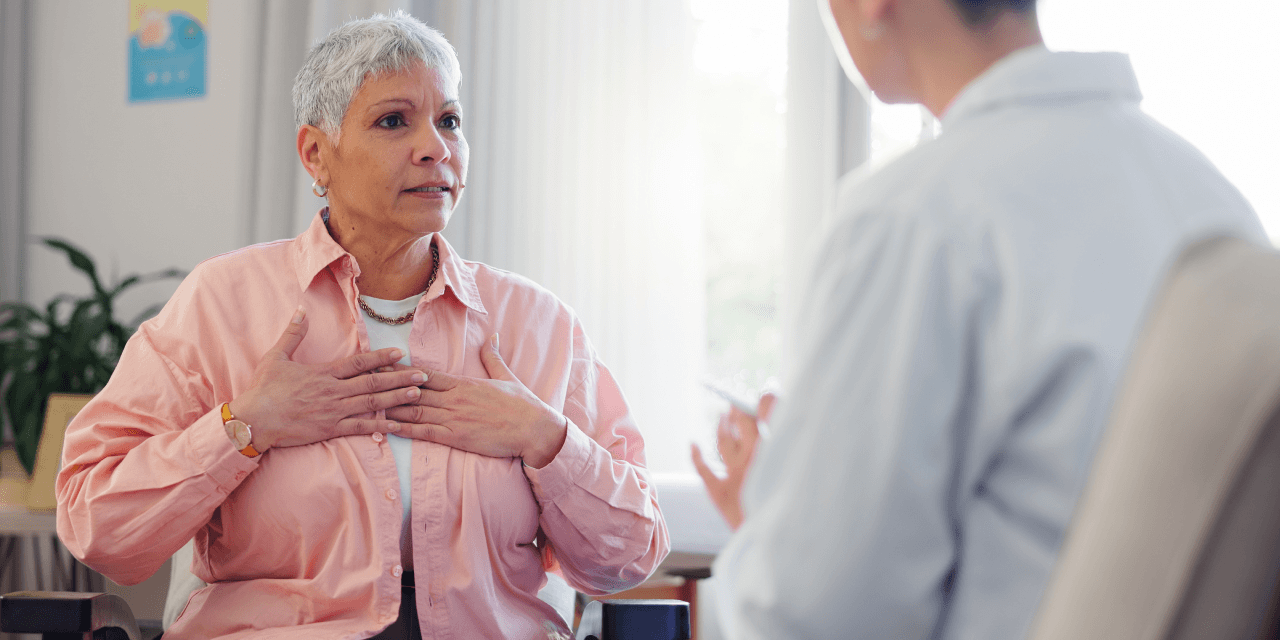 Older woman talking to her GP, calling attention to her chest area.