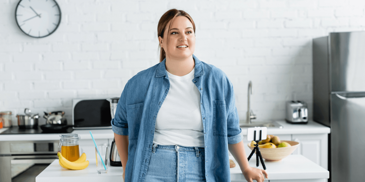 Young woman in denim shirt smiling. She's resting on a counter top against the airy, luminous backdrop of a modern kitchen.