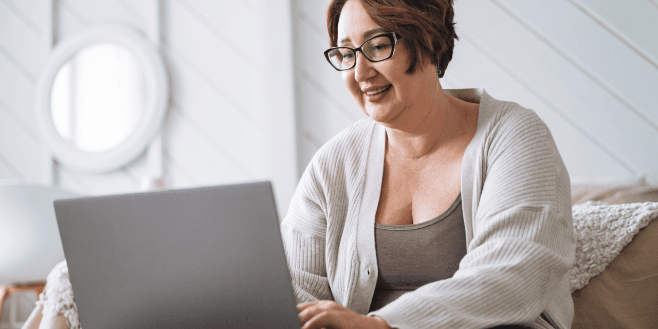 Middle-aged woman in beige cardigan is sitting on a sofa, using her laptop to research whether she can restart weight loss medication after stopping. She's in a modern, light, and airy living room, with a round mirror visible in the background.