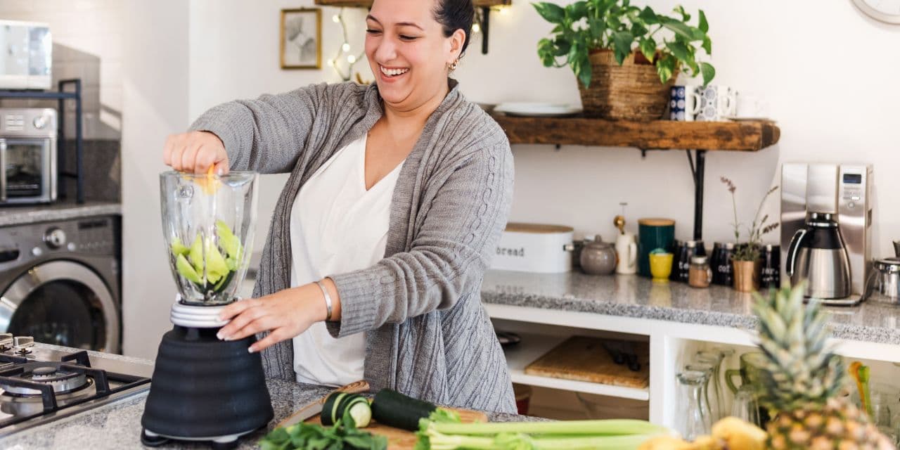 Woman in grey cardigan making a green smoothie in a modern kitchen.
